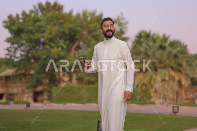 رجل عربي خليجي سعودي يتجول في احدى الحدائق العامة، استخدام الهاتف المحمول، المشي والتنزه، انشطة ترفيهية، الحدائق والمنتزهات، نزهه في الهواء الطلق