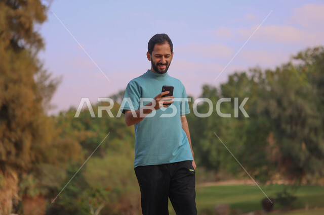 رجل عربي خليجي سعودي يتجول في احدى الحدائق العامة، استخدام الهاتف المحمول، المشي والتنزه، انشطة ترفيهية، الحدائق والمنتزهات، نزهه في الهواء الطلق