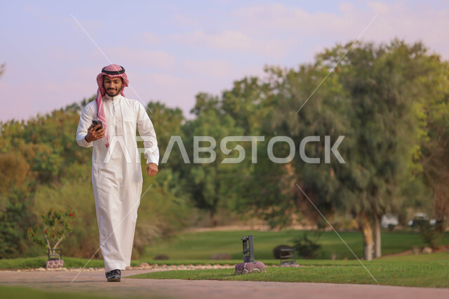 رجل عربي خليجي سعودي يتجول في احدى الحدائق العامة، استخدام الهاتف المحمول، المشي والتنزه، انشطة ترفيهية، الحدائق والمنتزهات، نزهه في الهواء الطلق