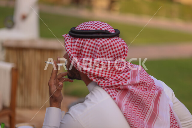 صورة مقربة لرجل عربي سعودي خليجي يقوم بتدخين السجائر، الجلوس في الهواء الطلق، الادمان على التبغ والسجائر، الطبيعة الخضراء