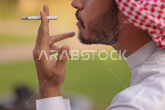 صورة مقربة لرجل عربي سعودي خليجي يقوم بتدخين السجائر، الجلوس في الهواء الطلق، الادمان على التبغ والسجائر، الطبيعة الخضراء
