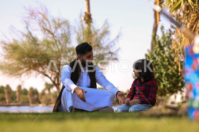 رجل عربي خليجي سعودي يقضي أوقات ممتعه مع ابنه، الجلوس في إحدى الحدائق العامة، دردشة ومحادثات, نزهه في الهواء الطلق،  الطبيعة الخضراء، أنشطة ترفيهية عائلية, عطله صيفيه