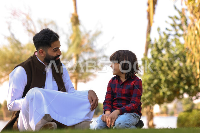 A Saudi Arabian Gulf man spending fun times with his son, sitting in a public park, chatting and talking, picnicking outdoors, green nature, family leisure activities, summer vacation