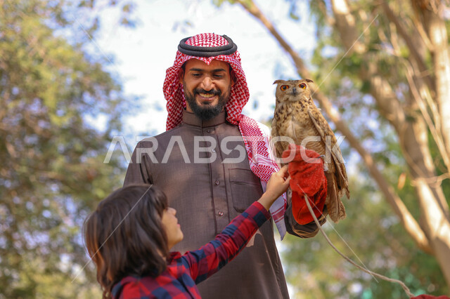 رجل عربي خليجي سعودي مع إبنه، قضاء أوقات ممتعة في حديقة الحيوانات في المملكة العربية السعودية، أماكن سياحية ترفيهية، أنشطة ترفيهية مع الطيور، تربية وتدريب الطيور الجارحة في المحمية الطبيعية، طائر البومة