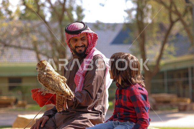 رجل عربي خليجي سعودي مع إبنه، قضاء أوقات ممتعة في حديقة الحيوانات في المملكة العربية السعودية، أماكن سياحية ترفيهية، أنشطة ترفيهية مع الطيور، تربية وتدريب الطيور الجارحة في المحمية الطبيعية، طائر البومة