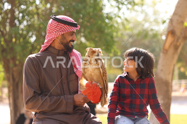 رجل عربي خليجي سعودي مع إبنه، قضاء أوقات ممتعة في حديقة الحيوانات في المملكة العربية السعودية، أماكن سياحية ترفيهية، أنشطة ترفيهية مع الطيور، تربية وتدريب الطيور الجارحة في المحمية الطبيعية، طائر البومة