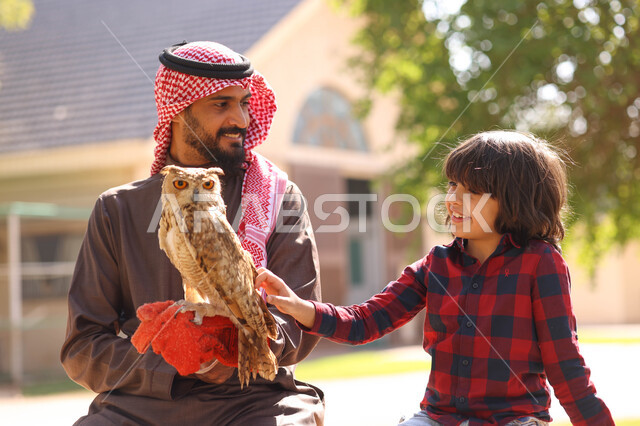 رجل عربي خليجي سعودي مع إبنه، قضاء أوقات ممتعة في حديقة الحيوانات في المملكة العربية السعودية، أماكن سياحية ترفيهية، أنشطة ترفيهية مع الطيور، تربية وتدريب الطيور الجارحة في المحمية الطبيعية، طائر البومة