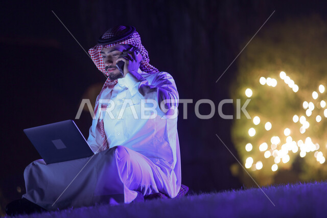 رجل عربي خليجي سعودي يستخدم الحاسوب المحمول، اجراء مكالمة هاتفية، تصفح الإنترنت، العمل عن بعد، عقد إجتماع عمل، هاتف محمول، استخدام تقنيات حديثة، حدائق ومنتزهات
