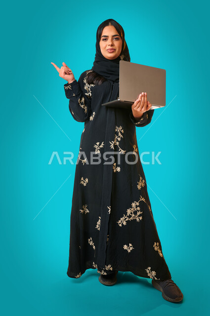 بورتريه لمرأة عربية خليجية سعودية ، إستخدام جهاز الحاسوب المحمول ، صورة بالطول الكامل , التعليم و العمل عن بعد ، اشارة باليد لمكان ما ، التسويق الإلكتروني ، تصفح الإنترنت ، مهن ووظائف  ، خلفية زرقاء .