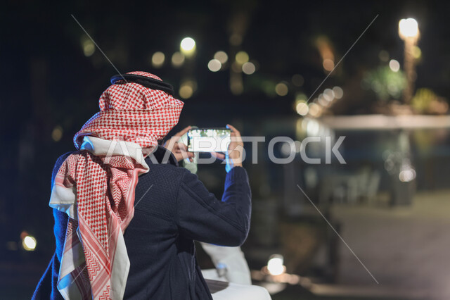 صورة من الخلف لرجل عربي خليجي سعودي يستخدم الهاتف المحمول المحمول، استخدام وسائل التواصل الاجتماعي، التقاط الصور من خلال الهاتف المحمول، كاميرا خلفية، بث مباشر، حدائق ومنتزهات