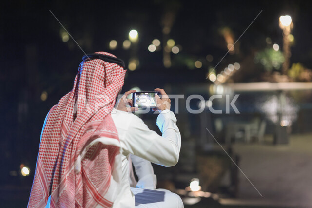 A picture from the back of a Saudi Arabian Gulf man using a mobile phone, using social media, taking pictures through a mobile phone, a rear camera, live broadcast, gardens and parks