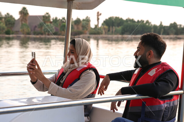 زوجان عربيان خليجيان سعوديان بقضيان اوقات ممتعة في احد الاماكن السياحية الترفيهية، ركوب قارب في بحيرة مائية، استخدام الهاتف المحمول، التقاط صور سيلفي تذكارية، بث مباشر، بركة مائية، نزهه في الهواء الطلق، أنشطة ترفيهية، أجواء عائلية سعيدة