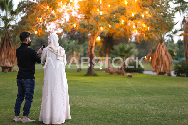 صورة من الخلف لزوجان عربيان خليجيان سعوديان يقضيان اوقات ممتعة في احد الحدائق السعودية، استخدام الهاتف المحمول، التقاط صور تذكارية، بث مباشر، نزهة في الهواء الطلق