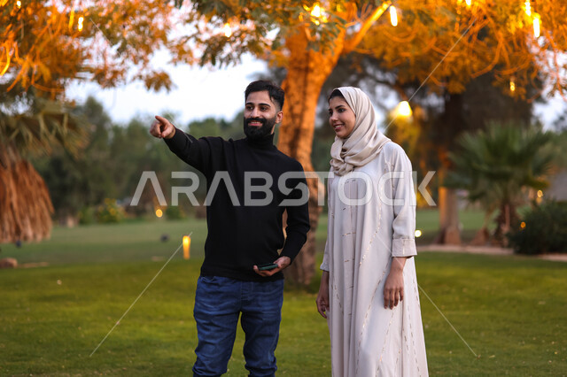 An Arab Gulf couple spending enjoyable times in a public park in Saudi Arabia, parks and gardens, green nature, pointing towards something, a happy family atmosphere, a picnic in the open air