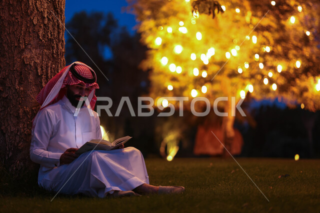 A Saudi Arabian Gulf man spending pleasant times in a public park, reading stories and novels, enjoying reading books, educating and acquiring knowledge, sitting outdoors