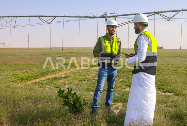 Two Saudi Arab Gulf agricultural engineers in an agricultural field, wearing a helmet and protection jacket, business agreements and deals, agricultural lands and fields, machines and equipment for future irrigation, pivot irrigation arms