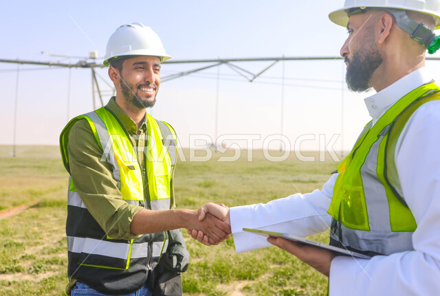Two Saudi Arab Gulf agricultural engineers in an agricultural field, wearing a helmet and protection jacket, business agreements and deals, agricultural lands and fields, machines and equipment for future irrigation, pivot irrigation arms