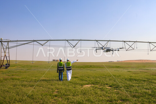 Two Saudi Arab Gulf agricultural engineers in an agricultural field, wearing a helmet and protection jacket, business agreements and deals, agricultural lands and fields, machines and equipment for future irrigation, pivot irrigation arms