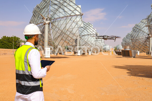 مهندس عربي خليجي سعودي، يرتدي الخوذة وسترة الحماية، يحمل بيده جهاز تقني، التدقيق والإشراف على العمل، مفهوم هندسة الطاقة، مهن ووظائف سعودية، محطات الطاقة الشمسية، ألياف شمسية، ستلايت