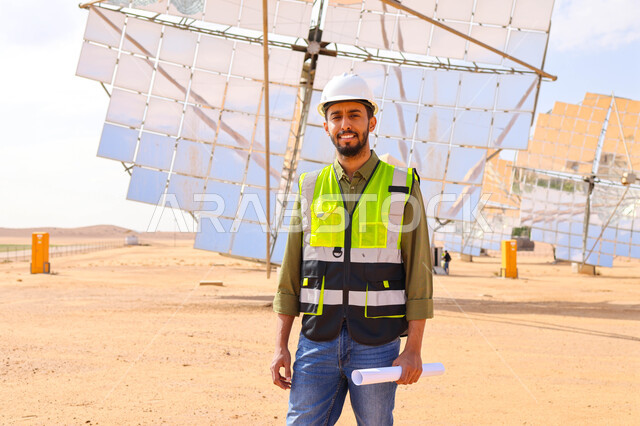مهندس عربي خليجي سعودي، يرتدي الخوذة وسترة الحماية، يحمل بيده مخططات وتصاميم هندسية، التدقيق والإشراف على العمل، مفهوم هندسة الطاقة، مهن ووظائف سعودية، محطات الطاقة الشمسية، ألياف شمسية، ستلايت