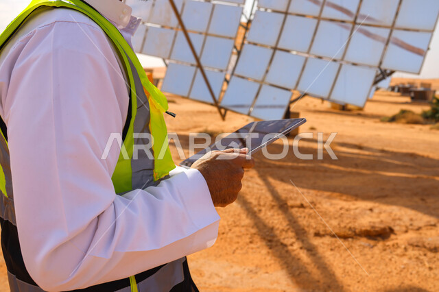 مهندس عربي خليجي سعودي، يرتدي الخوذة وسترة الحماية، يحمل بيده جهاز تقني، التدقيق والإشراف على العمل، مفهوم هندسة الطاقة، مهن ووظائف سعودية، محطات الطاقة الشمسية، ألياف شمسية، ستلايت