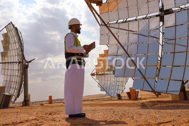 مهندس عربي خليجي سعودي، يرتدي الخوذة وسترة الحماية، يحمل بيده جهاز تقني، التدقيق والإشراف على العمل، مفهوم هندسة الطاقة، مهن ووظائف سعودية، محطات الطاقة الشمسية، ألياف شمسية، ستلايت
