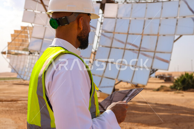 مهندس عربي خليجي سعودي، يرتدي الخوذة وسترة الحماية، يحمل بيده جهاز تقني، التدقيق والإشراف على العمل، مفهوم هندسة الطاقة، مهن ووظائف سعودية، محطات الطاقة الشمسية، ألياف شمسية، ستلايت