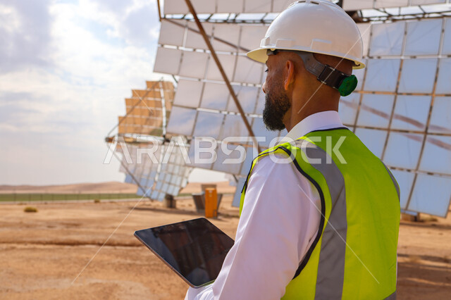 مهندس عربي خليجي سعودي، يرتدي الخوذة وسترة الحماية، يحمل بيده جهاز تقني، التدقيق والإشراف على العمل، مفهوم هندسة الطاقة، مهن ووظائف سعودية، محطات الطاقة الشمسية، ألياف شمسية، ستلايت