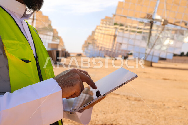 مهندس عربي خليجي سعودي، يرتدي الخوذة وسترة الحماية، يحمل بيده جهاز تقني، التدقيق والإشراف على العمل، مفهوم هندسة الطاقة، مهن ووظائف سعودية، محطات الطاقة الشمسية، ألياف شمسية، ستلايت