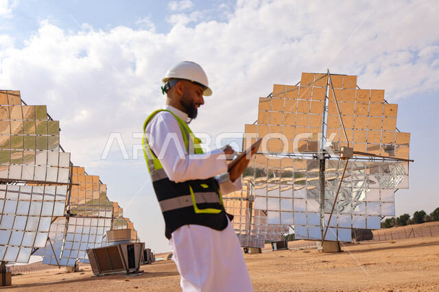 مهندس عربي خليجي سعودي، يرتدي الخوذة وسترة الحماية، يحمل بيده جهاز تقني، التدقيق والإشراف على العمل، مفهوم هندسة الطاقة، مهن ووظائف سعودية، محطات الطاقة الشمسية، ألياف شمسية، ستلايت