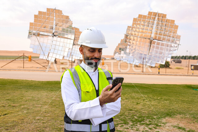 مهندس عربي خليجي سعودي، يرتدي الخوذة وسترة الحماية، يحمل بيده الهاتف المحمول، التدقيق والإشراف على العمل، مفهوم هندسة الطاقة، مهن ووظائف سعودية، محطات الطاقة الشمسية، ألياف شمسية، ستلايت