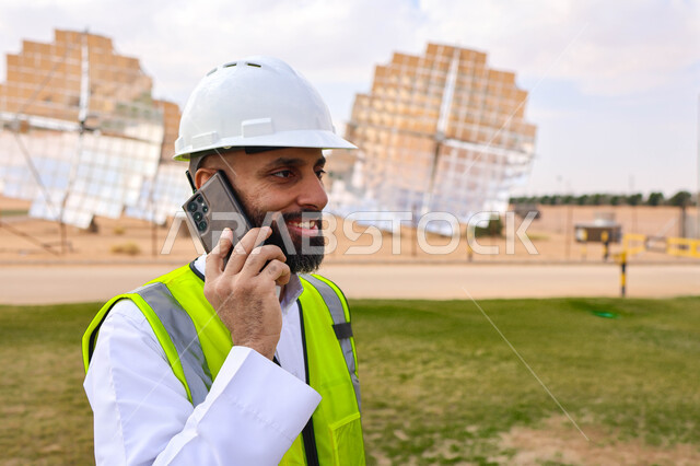 مهندس عربي خليجي سعودي، يرتدي الخوذة وسترة الحماية، يحمل بيده الهاتف المحمول، التدقيق والإشراف على العمل، مفهوم هندسة الطاقة، مهن ووظائف سعودية، محطات الطاقة الشمسية، ألياف شمسية، ستلايت