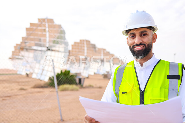مهندس عربي خليجي سعودي، يرتدي الخوذة وسترة الحماية، يحمل بيده مخططات وتصاميم هندسية، التدقيق والإشراف على العمل، مفهوم هندسة الطاقة، مهن ووظائف سعودية، محطات الطاقة الشمسية، ألياف شمسية، ستلايت