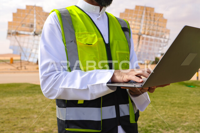 مهندس عربي خليجي سعودي، يرتدي الخوذة وسترة الحماية، يحمل بيده جهاز تقني، التدقيق والإشراف على العمل، مفهوم هندسة الطاقة، مهن ووظائف سعودية، محطات الطاقة الشمسية، ألياف شمسية، ستلايت