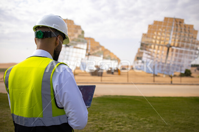 مهندس عربي خليجي سعودي، يرتدي الخوذة وسترة الحماية، يحمل بيده جهاز تقني، التدقيق والإشراف على العمل، مفهوم هندسة الطاقة، مهن ووظائف سعودية، محطات الطاقة الشمسية، ألياف شمسية، ستلايت