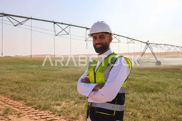 مهندس زراعي عربي خليجي سعودي يقف في حقل زراعي، إرتداء خوذة وسترة الحماية ، الإشراف على العمل, الات ومعدات الري المستقبلي، اذرع الري المحوري