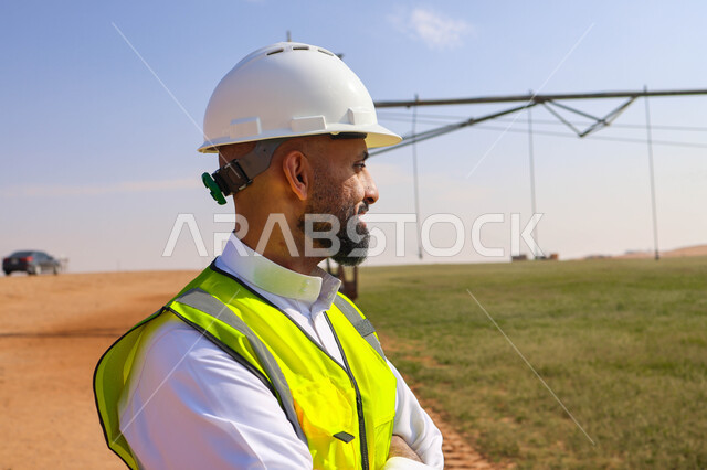 مهندس زراعي عربي خليجي سعودي يقف في حقل زراعي، إرتداء خوذة وسترة الحماية ، الإشراف على العمل, الات ومعدات الري المستقبلي، اذرع الري المحوري
