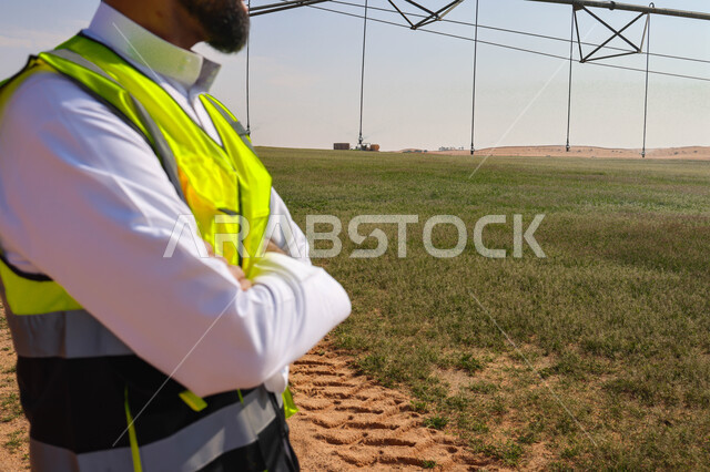 مهندس زراعي عربي خليجي سعودي يقف في حقل زراعي، إرتداء خوذة وسترة الحماية ، الإشراف على العمل, الات ومعدات الري المستقبلي، اذرع الري المحوري