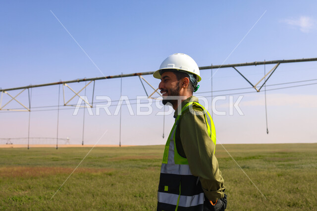 مهندس زراعي عربي خليجي سعودي يقف في حقل زراعي، إرتداء خوذة وسترة الحماية ، الإشراف على العمل, الات ومعدات الري المستقبلي، اذرع الري المحوري