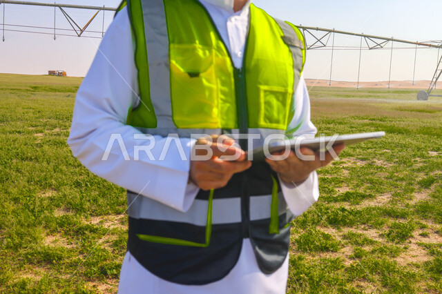 A close-up of a Saudi Arabian Gulf agricultural engineer standing in an agricultural field, holding a technical device in his hand, wearing a protection jacket, supervising work, future irrigation machines and equipment, pivot irrigation arms