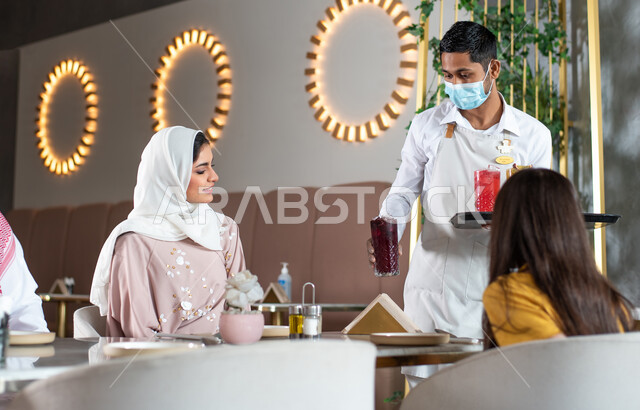 كافيهات ومطاعم في السعودية, جلسات فريدة ومميزة,  النادل يقوم بتقديم الحلويات والسكريات, زوجان عربيان سعوديان خليجيان يجلسان في المقهى, إيماءات تدل على السعادة, تناول المشروبات و قضاء وقت ممتع في الخارج مع العائلة, دردشة ومحادثات