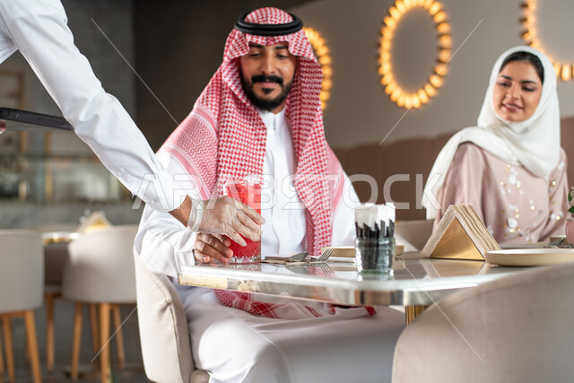 كافيهات ومطاعم في السعودية, النادل يقوم بتقديم الحلويات والسكريات, زوجان عربيان سعوديان خليجيان يجلسان في المقهى, إيماءات تدل على السعادة, تناول المشروبات و قضاء وقت ممتع في الخارج مع العائلة, دردشة ومحادثات