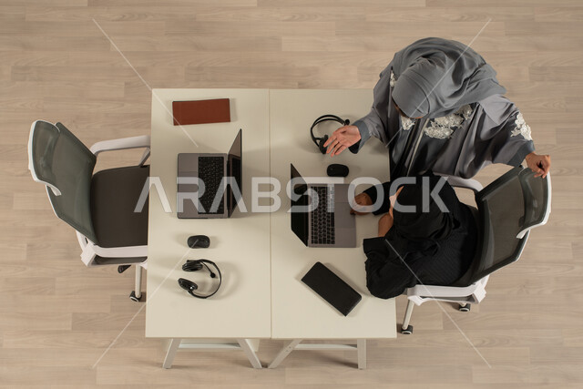 تقديم الاستشارات ومتابعة العمل، صورة عامودية علوية لموظفة عربية خليجية سعودية تستخدم الحاسوب المحمول وتقف بجانبها موظفة، العمل في الدعم الفني وخدمة العملاء، سماعات رأس صوتية، استعلامات واستفسارات، متابعة وتطوير العمل