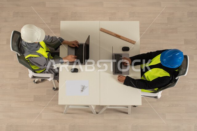 Wearing the work jacket and protection helmet, an upper vertical image of two Saudi Gulf engineers, completing work tasks through a laptop computer, the profession of architecture, Saudi employment and job opportunities, Saudi office professions and jobs