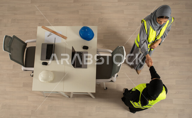 استخدام جهاز الحاسوب المحمول، العمالة السعودية وفرص العمل، صورة عامودية علوية لمهندستان خليجيتان سعوديتان تتصافحان، توقيع صفقات جديدة، اتفاقيات عمل ناجحة، ارتداء السترة الخاصة بالعمل، مهنة الهندسة المعمارية، مهن ووظائف مكتبية سعودية