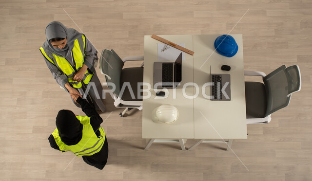 ارتداء السترة الخاصة بالعمل، استخدام جهاز الحاسوب المحمول، صورة عامودية علوية لمهندستان خليجيتان سعوديتان تتصافحان، توقيع صفقات جديدة، اتفاقيات عمل ناجحة، مهنة الهندسة المعمارية، العمالة السعودية وفرص العمل، مهن ووظائف مكتبية سعودية