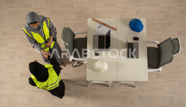 اتفاقيات عمل ناجحة، العمالة السعودية وفرص العمل، صورة عامودية علوية لمهندستان خليجيتان سعوديتان تتصافحان، توقيع صفقات جديدة، استخدام جهاز الحاسوب المحمول، ارتداء السترة الخاصة بالعمل، مهنة الهندسة المعمارية، مهن ووظائف مكتبية سعودية