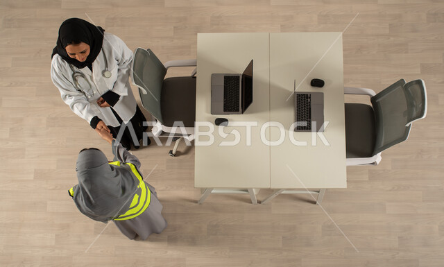 استخدام جهاز الحاسوب المحمول، صورة عامودية علوية لطبيبة ومهندسة معمارية خليجيتان سعوديتان تتصافحان، توقيع صفقات جديدة، اتفاقيات عمل ناجحة، العمالة السعودية وفرص العمل، عمل جماعي مشترك، مهن ووظائف مكتبية سعودية