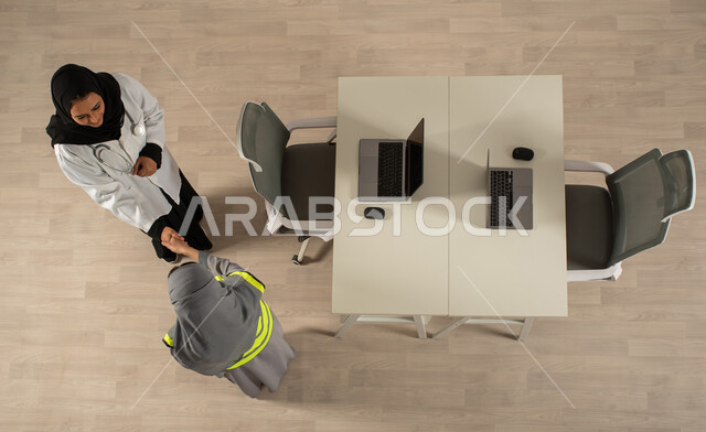 العمالة السعودية وفرص العمل، صورة عامودية علوية لطبيبة ومهندسة معمارية خليجيتان سعوديتان تتصافحان، استخدام جهاز الحاسوب المحمول، توقيع صفقات جديدة، اتفاقيات عمل ناجحة، عمل جماعي مشترك، مهن ووظائف مكتبية سعودية