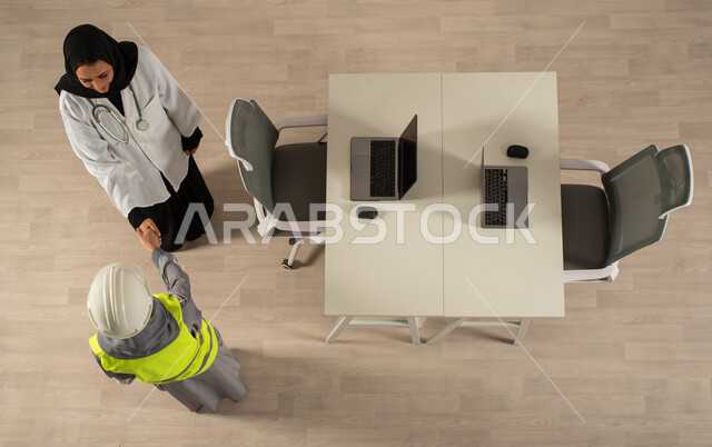 العمل الجماعي المشترك، صورة عامودية علوية لطبيبة ومهندسة معمارية خليجيتان سعوديتان تتصافحان، استخدام جهاز الحاسوب المحمول، توقيع صفقات جديدة، اتفاقيات عمل ناجحة، العمالة السعودية وفرص العمل، مهن ووظائف مكتبية سعودية
